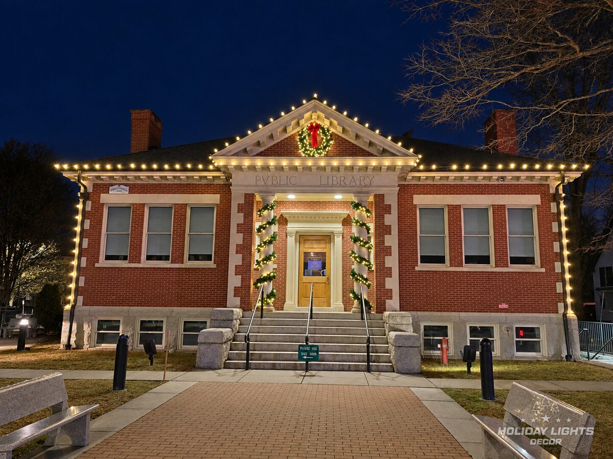 Brick civic building with warm-white roofline lights, a wreath, and garland-wrapped columns.