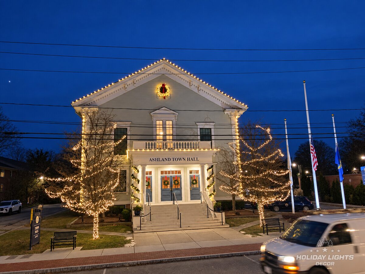 Civic building outlined in warm-white lights with lit trees, wreaths, and garland.