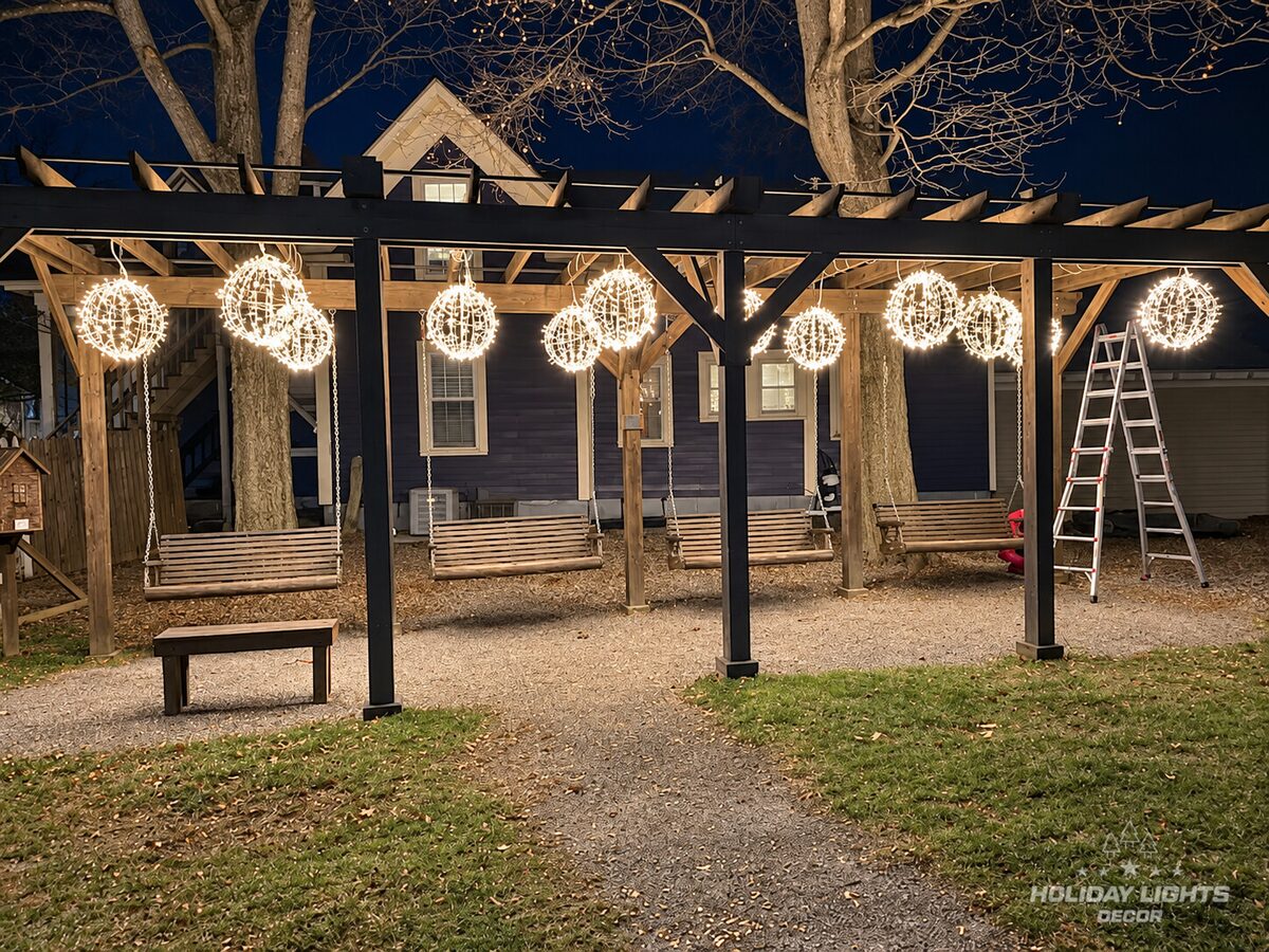 Pergola with illuminated spheres suspended from its beams above a gravel path.