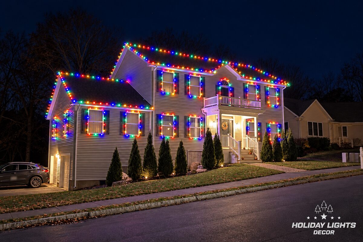 Nighttime street view of a two-story home with multicolor lights outlining the rooflines, windows, porch, and balcony.