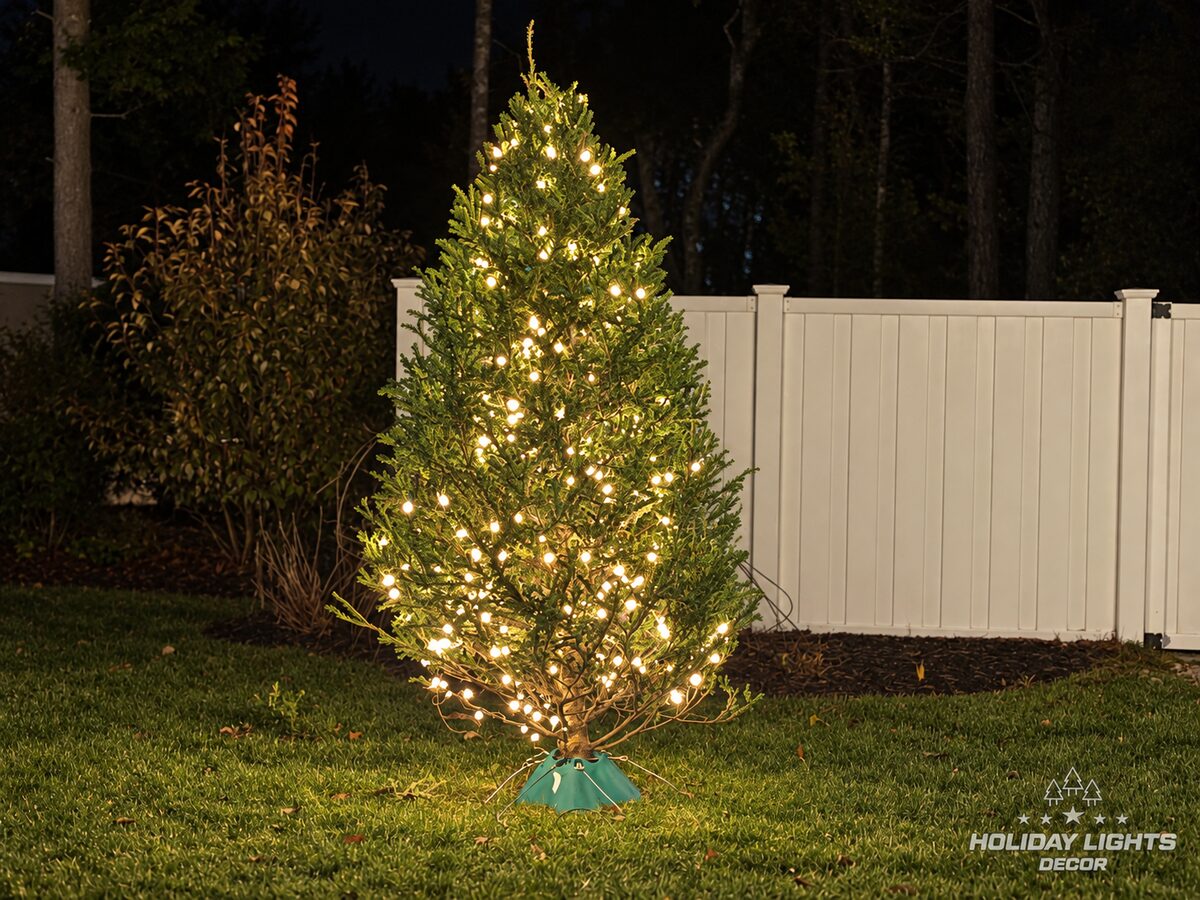 Small backyard tree wrapped in warm-white lights beside a white vinyl fence