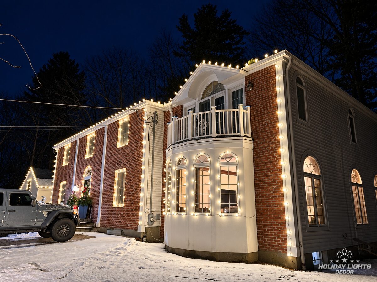 Side angle of a Lynnfield, MA brick home with warm white lights outlining the balcony gable, bay windows, corners and roofline in the snow