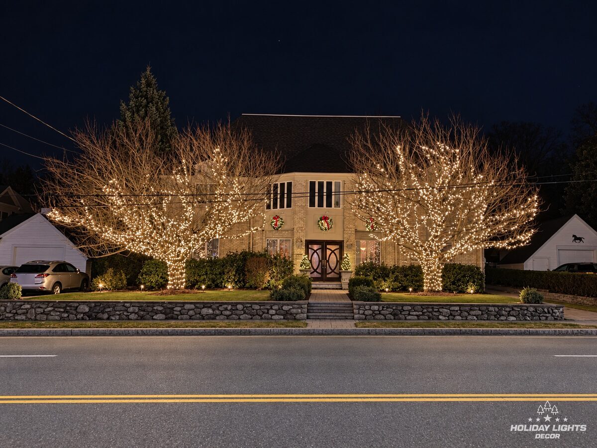 Street view of a Waltham, MA home with two large trees wrapped in warm white mini lights framing the entrance and wreaths above