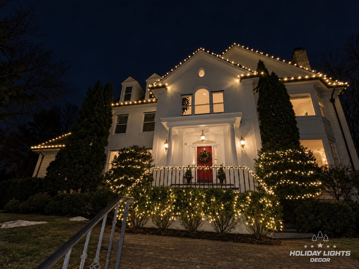 Front view of a white Brookline, MA estate with warm white C9 gables, lit shrubs, stair railing and a wreath on the red door