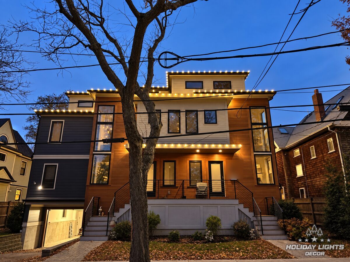 Front evening view of a modern Brookline, MA home with warm white lights on the upper roof edges and entrance canopy, framed by a mature tree