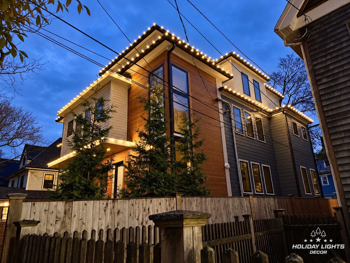 Side view of a modern Brookline, MA home with warm white lights across multiple roof levels above wood siding, tall windows and a fenced yard