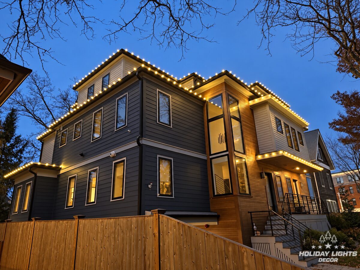 Angled evening view of a modern Brookline, MA home with warm white lights along the stepped rooflines, side eaves and entrance overhang