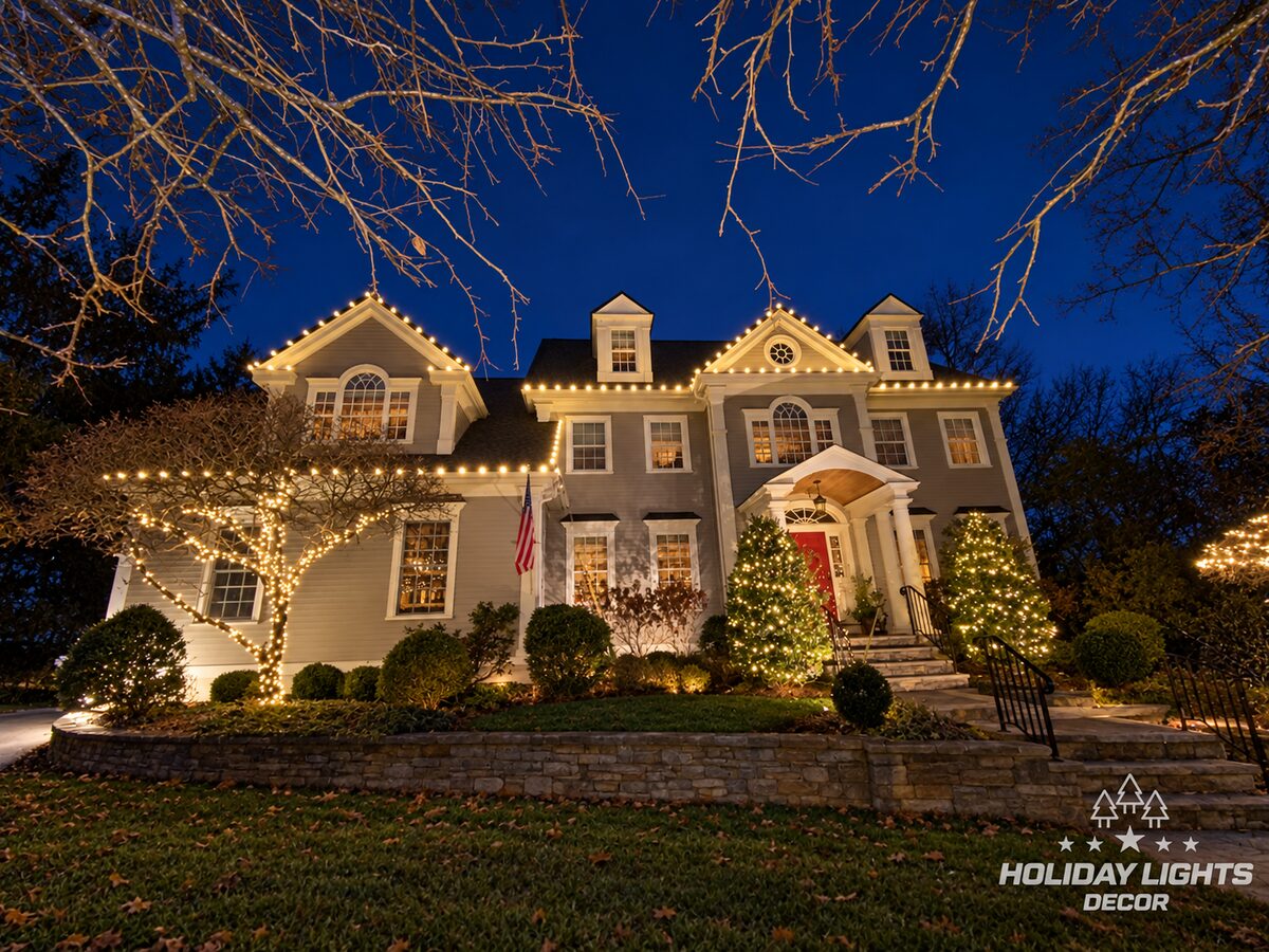 Closer view of a Lexington, MA colonial with roofline lights, a wrapped ornamental tree and lit evergreens beside the red front door
