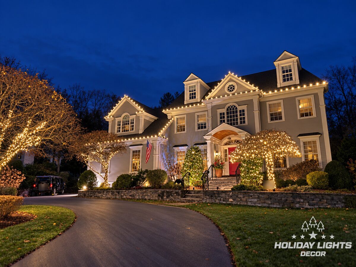 Angled view of a Lexington, MA colonial with warm white C9 lights on the front gables, a lit entrance and ornamental trees in mini lights